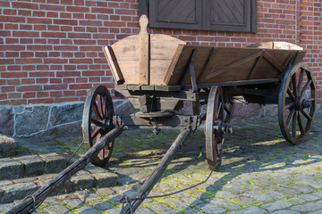 A brown wooden vintage cart with wooden wheels, standing drawbar forward on an old gray stone sidewalk overgrown with grass against a red brick wall with a shuttered window at the stone porch