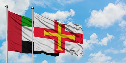 United Arab Emirates and Guernsey flag waving in the wind against white cloudy blue sky together....