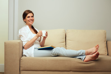 Smiling woman drinking coffee sitting on sofa with book.