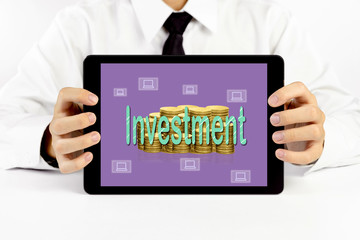 Businessman holding a tablet with green word Investment and golden coins stacks and laptop icons