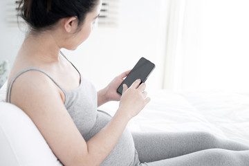 Fototapeta na wymiar Young pregnant woman using smartphone with blank screen lying on bed. Young expectant lady surfing net, reading about baby care, buying goods for newborn concept.