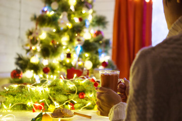 Famale hands holding a cozy ceramic handmade mug with coffe. Winter and Christmas home time concept. Lifestyle. Copy space Decorating and christmas tree