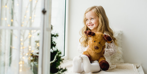 little girl with a plush deer sitting on the window. A child looks out the window and is waiting for Christmas, Santa Claus