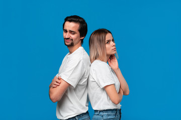 Upset couple of a young blond woman and brunet bearded man with mustaches in white t-shirts and blue jeans posing isolated over blue background. Concept of controversy