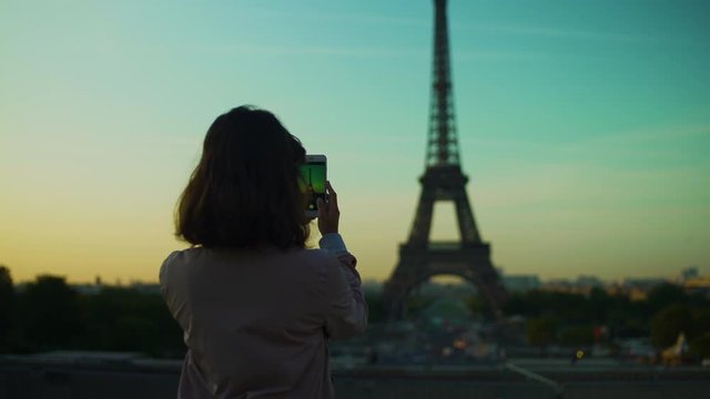 Girl taking picture with smartphone of Eiffel tower at sunset orange blue sky in Paris during the summer. Haussmanian buildings, trees, 16th, symbol, trocadero. Side track steadicam 4K UHD.