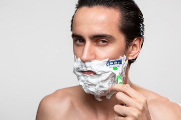 Handsome man with mustache uses shaving stick and shaving foam to trim his beard standning bare...