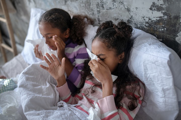 Two sisters lying in bed and sneezing after catching flu - obrazy, fototapety, plakaty