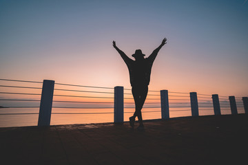 Fototapeta premium Silhouette of one lonely free carefree traveler woman in hat with open outstretched arms standing on sea pier at sunset. Happy freedom concept and enjoying life
