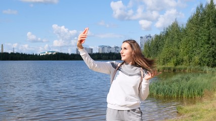 Beautiful girl with a backpack takes a selfie near the lake and talks on a cell phone on Skype.