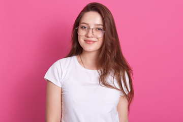 Relaxed carefree smiling young woman wearing white casual t shirt and glasses, having positive facial expression, looking directly at camera. Horizontal studio shot isolated over pink background.