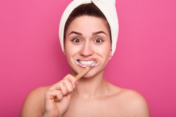 Horizontalshot of adorable woman brushing her teeth, having astonish facial expression, posing with white towel on her head, standing with wide opened eyes and bared shoulders. Oral hygiene concept.