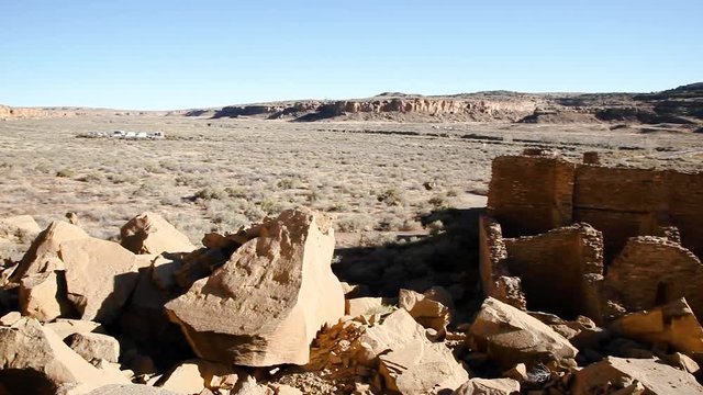 Panning across an overlook of Pueblo Bonito at Chaco Canyon in New Mexico. It is late afternoon and the sun flares in the lens for dramatic effect halfway through. Two unidentifiable people walk below