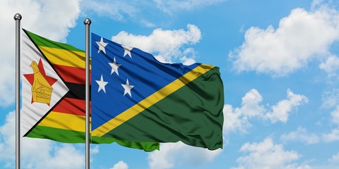 Zimbabwe and Solomon Islands flag waving in the wind against white cloudy blue sky together....