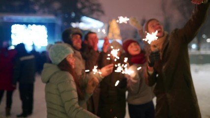 Friends have fun partying with sparklers and doing selfie photo on smartphone at Christmas market. People jump and dance at New Year fairground on winter night. Snowfall, bulb garlands on backdrop. - Powered by Adobe