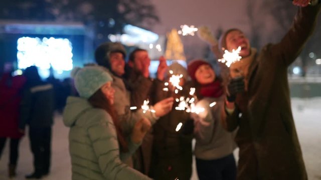 Friends have fun partying with sparklers and doing selfie photo on smartphone at Christmas market. People jump and dance at New Year fairground on winter night. Snowfall, bulb garlands on backdrop.