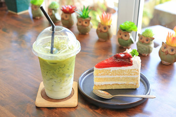 Strawberry cake in metal tray and Iced matcha green tea latte in take away cup on wooden table at cafe.