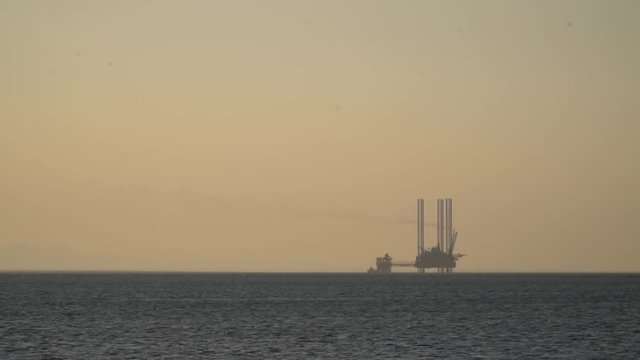 Silhouette of a massive oil rig in the sea, orange foggy background, sunset