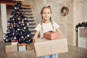 Obraz na płótnie Canvas Portrait of pretty little girl holding her Christmas presents and smiling at camera while standing in the hall with Christmas tree in the background