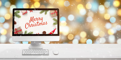 Merry Christmas greeting on computer display on white wooden desk. Bokeh and Christmas lights in background. Copy space beside. Keyboard and mouse on desk.