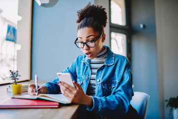 Shocked female student in jeans jacket surprised of receiving email with discounts during learning with knowledge textbook indoors, amazed hipster girl reading news about buscary on university website