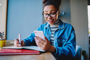 Happy hipster girl in eyeglasses feeling excited of new notification on modern cellphone while...