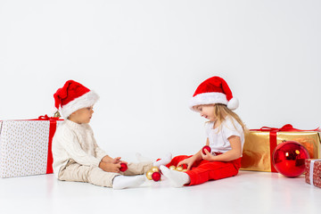 Funny little kids in Santa hat sitting between gift boxes and playing with christmas balls. Isolated on white background. New year