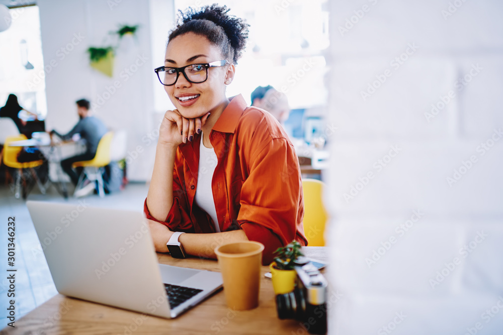 Poster portrait of positive hipster girl in optical eyewear looking at camera during using laptop computer 