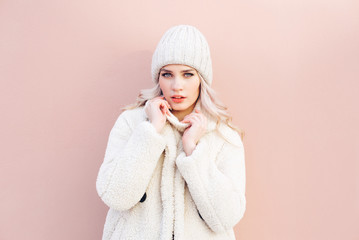 Happy blonde girl in white winter clothes posing against a pink wall.