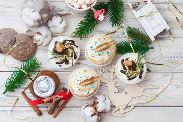 Cupcakes with cream cheese frosting and tangerines, cotton flowers, christmas tree branches, mini wooden sledge, chocolate cookies, caramel jar with red riboon, cinnamon rolls on wooden background.