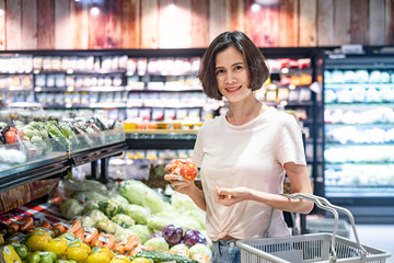 Young Asian beautiful woman holding grocery basket walking in supermarket. She is holding tomato in vegetable and fruit area with smile. Seen from front while she looking on camera. Healthy shopping.