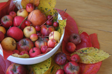 Little red and yellow apples in a white bowl on wooden table with red and yellow leaves. Autumn background