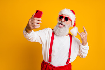 Fototapeta na wymiar X-mas party hard. Portrait of funny crazy santa claus in red hat take selfie smart phone christmas blogger show horns grimace tongue out wear shirt suspenders isolated yellow color background