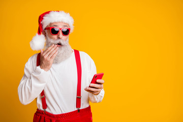 Fototapeta na wymiar Omg newyear x-mas discounts. Portrait of impressed funky crazy grey hair santa claus in red hat use smart phone read noel christmas information isolated yellow color background