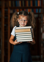 Cute little girl in glasses stands in the library full of books. Conception of education