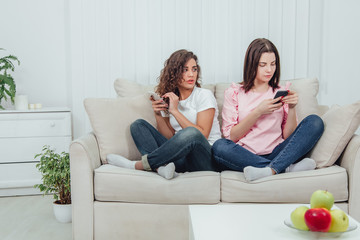 Two attractive girlfriends sitting on the sofa side by side in lotos position, with smartphones in...