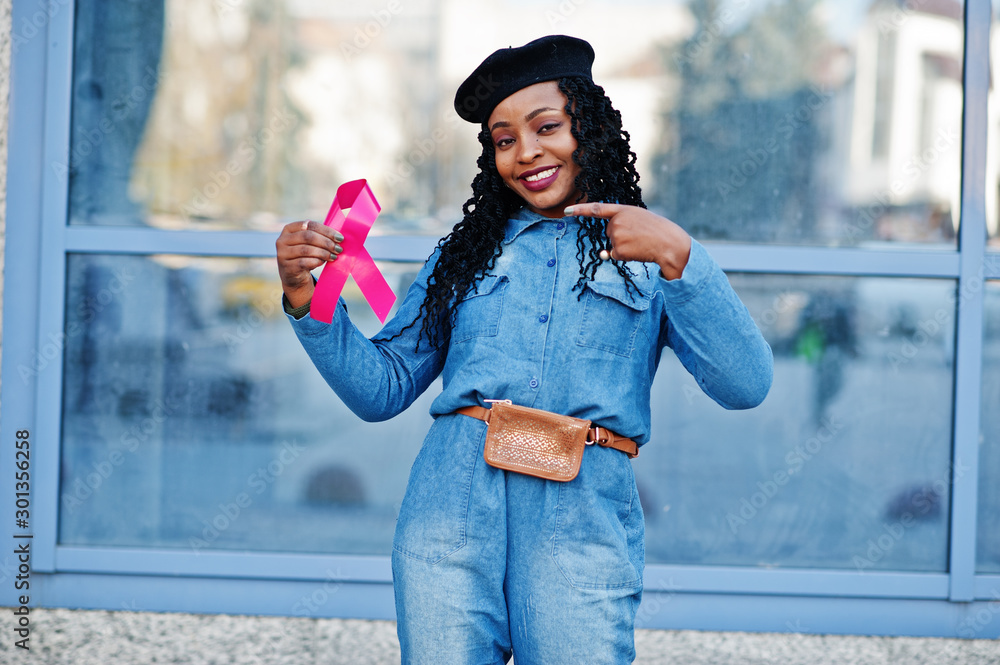 Wall mural The power to fight. Stylish fashionable african american women in jeans wear and black beret against modern building with pink ribbon for breast cancer.