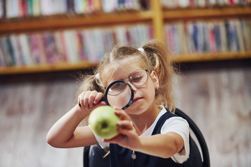 Holds magnifier. Cute little girl with pigtails is in the library. Apple on the books