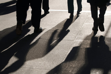 Morning sun casting dramatic shadows on the sidewalk behind silhouettes of commuting business people rushing to work