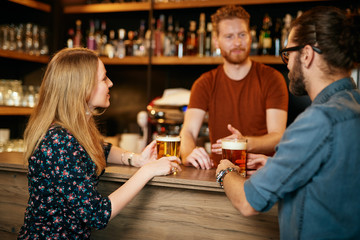 Cheerful friends leaning on bar counter, drinking beer and chatting with bartender. Night out.