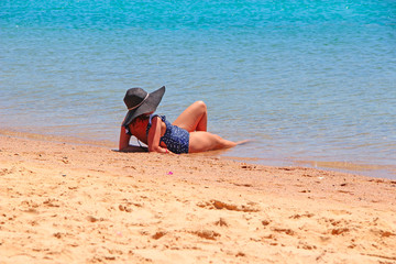 Fototapeta na wymiar Modern woman in dark hat lying by sea on beach