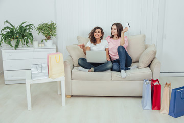 Obraz premium Two happy female friends using laptop for shopping online. One girl is holding credit card, another pointing her finger at numerous spop bags all around the room.