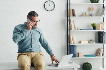 handsome businessman in shirt and glasses sitting on table and talking on smartphone