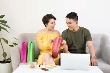 Young Couple Shopping Online On Laptop With Multi Colored Shopping Bags On Sofa