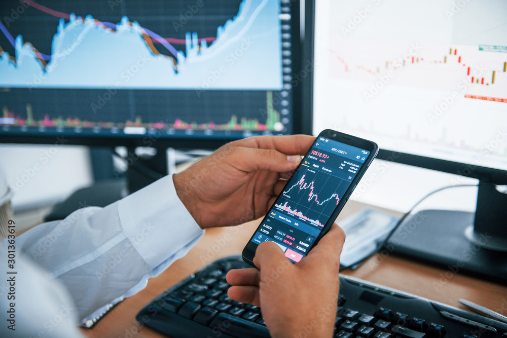 Poster close up view of phone holds by man's hands. businessman is in office with multiple screens. concept