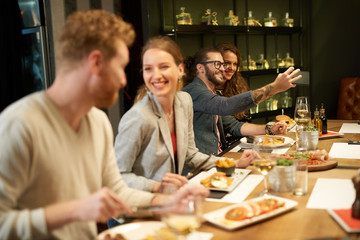 Cute caucasian fashionable couple sitting in restaurant at dinner and taking selfie. In foreground are their friends eating diner.
