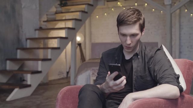 Young man using a smartphone sitting on the armchair at home in duplex apartment with garland lights behind him. Stock footage. Male with cell phone relaxing in the spacious room.