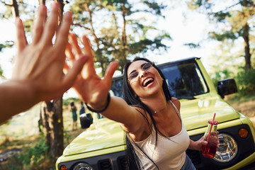 Gives high five. Blonde with alcohol in hand have a walk in the forest against green jeep - obrazy, fototapety, plakaty