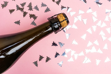 The neck of a bottle of champagne on a pink background with confetti in the shape of Christmas...
