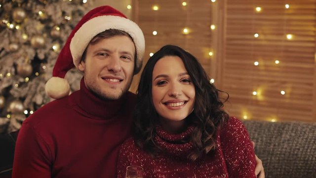 Portrait of Happy Smiling Couple in love Sitting near Christmas Tree at Warm and Cozy Home Atmosphere Holding Glasses of Chamagne. Concept of Holidays and New Year.