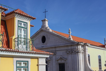 Fototapeta na wymiar The beautiful facade of a richly finished yellow house on the large glass doors and windows, the detailed iron balconies next to a colonial-style mud-roofed church in the city of Lisbon, Portugal.
